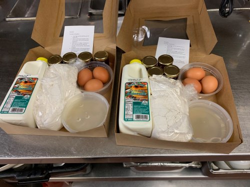 Two takeout boxes containing milk, eggs, flour, and jars, with recipe cards on a kitchen counter.