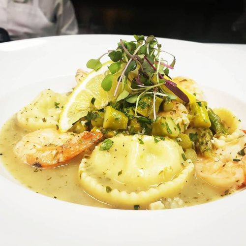 A plate of ravioli topped with shrimp, asparagus, and microgreens, garnished with a lemon slice in a creamy sauce.
