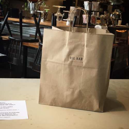 A brown paper bag from Pie Bar sits on a table in a café, with chairs and decor in the background.