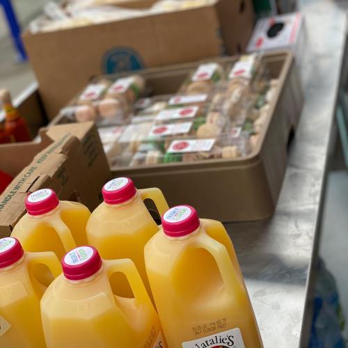 Bottles of orange juice in the foreground with packaged food items in the background on a stainless steel surface.