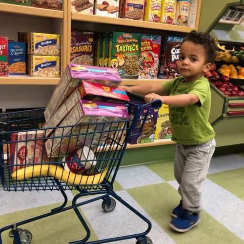 A young child pushes a shopping cart filled with cereal boxes and a banana in a grocery store aisle.