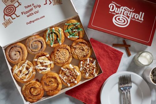 A box of assorted cinnamon rolls with various toppings, alongside a red napkin, fork, and glass of milk.