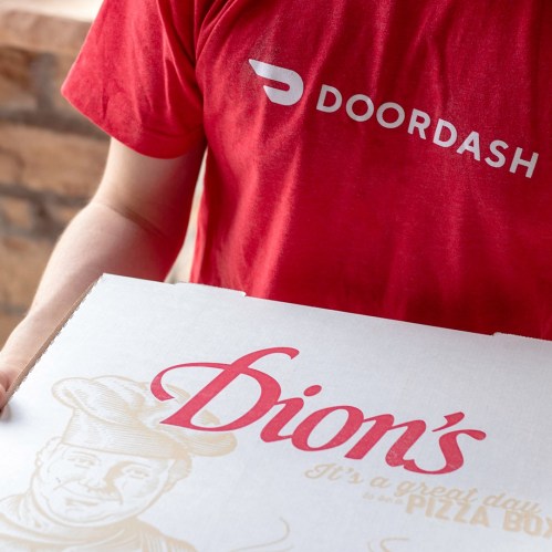 A person in a red DoorDash shirt holds a large Dion's pizza box with a chef illustration and slogan.