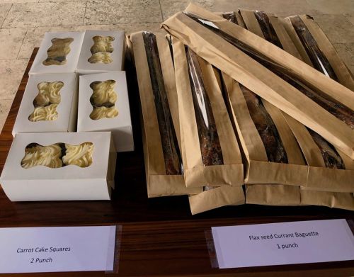 A wooden table displays packaged carrot cake squares and flax seed currant baguettes, labeled with their prices.