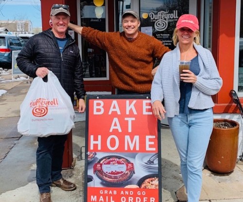 Three people stand outside a bakery, smiling and posing with a takeout bag and a drink, next to a "Bake at Home" sign.