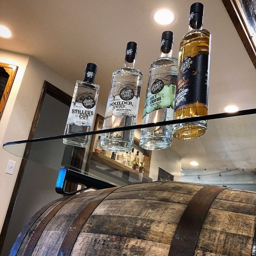Four bottles of spirits displayed on a glass shelf above a wooden barrel in a well-lit room.