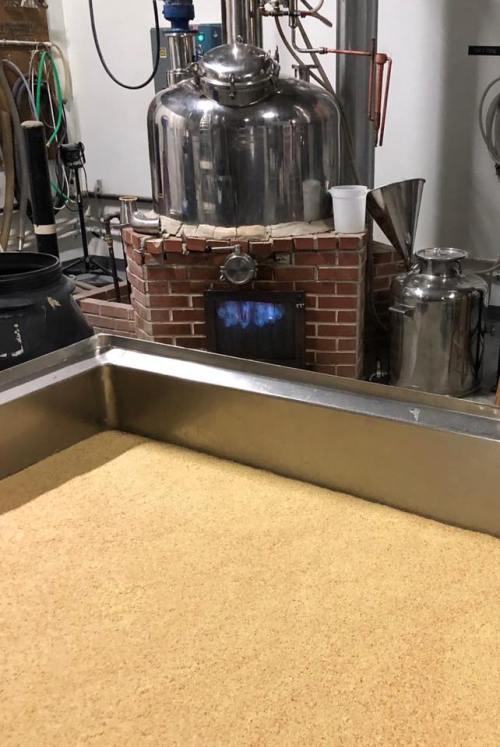 A distillery setup featuring a large metal still, a brick fireplace, and a tray of grain mash in the foreground.