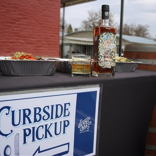 A table with a "Curbside Pickup" sign, featuring food containers and a bottle of whiskey with a glass beside it.