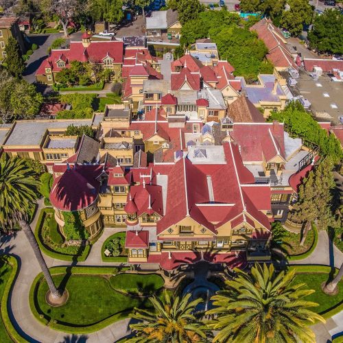 Aerial view of a large, ornate mansion with red roofs, surrounded by lush greenery and palm trees.