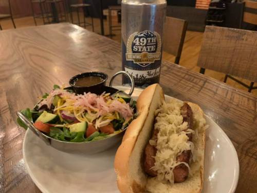 A plate with a hot dog topped with sauerkraut, a side salad, and a can of 49th State Brewing Company beer.