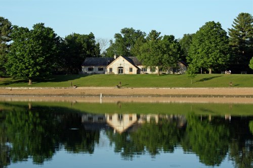 A serene lake reflects a stone house surrounded by lush green trees and a sandy shore under a clear blue sky.