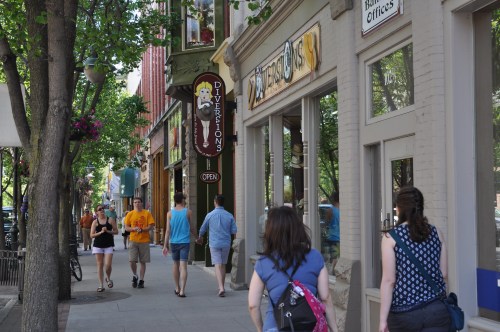 A busy street scene with people walking past shops and trees lining the sidewalk on a sunny day.