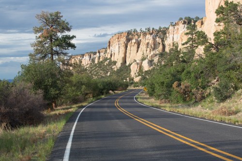 A winding road curves alongside rocky cliffs and lush greenery under a cloudy sky.