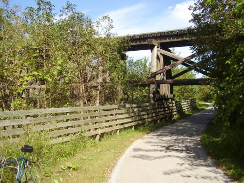 A bike path surrounded by greenery, with a wooden bridge overhead and a bicycle parked nearby.