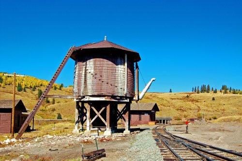 A weathered wooden water tower stands beside railway tracks in a rural landscape under a clear blue sky.