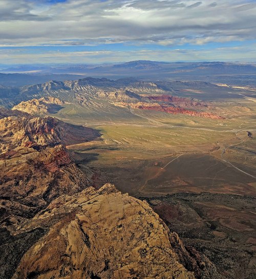 A panoramic view of rugged mountains and valleys under a cloudy sky, showcasing diverse terrain and colors.