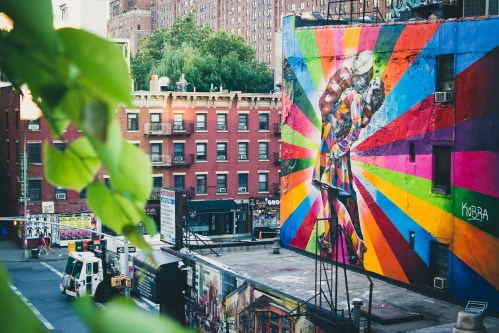 A vibrant mural of a colorful figure against a rainbow background, with urban buildings in the background.