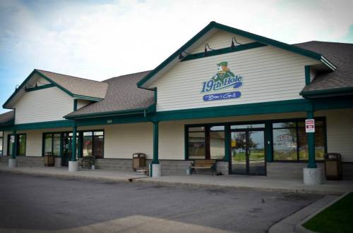 Exterior of a building with a sign reading "19th Hole" and a green roof, featuring benches and large windows.