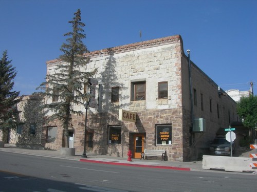 Historic stone building with a café sign, surrounded by trees and a clear blue sky. Street view with a bench nearby.