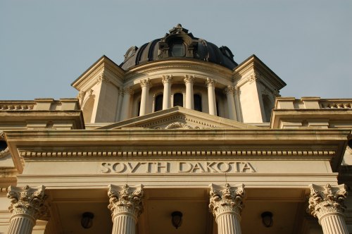 The ornate facade of the South Dakota State Capitol building, featuring columns and a dome against a clear sky.