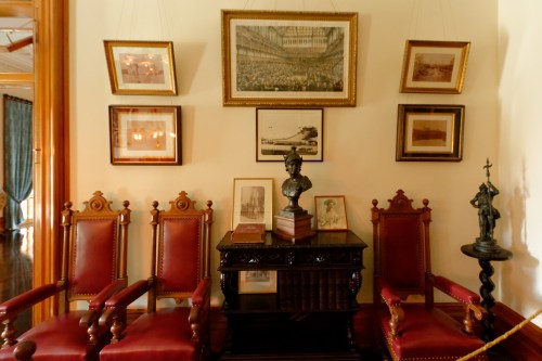 A vintage room featuring red upholstered chairs, a wooden table with books, and framed historical artwork on the walls.
