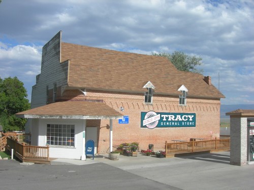A brick building with a sign reading "Tracy General Store," featuring a wooden deck and blue mailbox outside.