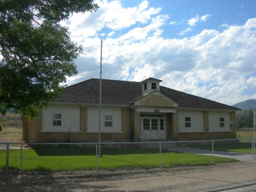 A brick building with a sloped roof, surrounded by grass and a fence, under a partly cloudy sky.