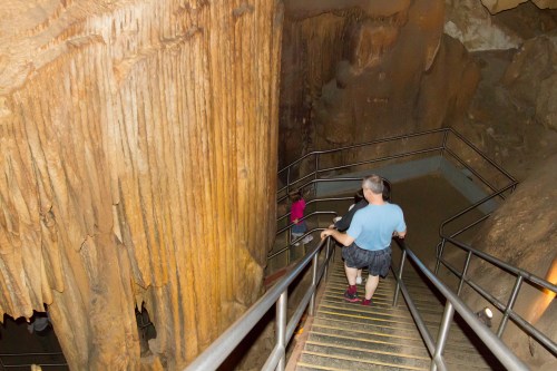 A person descends metal stairs into a cave, surrounded by large, textured rock formations.