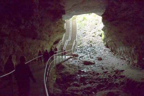 A dimly lit cave entrance with a winding path and stairs leading up, surrounded by rocky terrain and greenery.