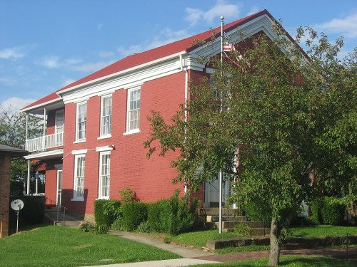 A red brick building with a white porch and an American flag, surrounded by greenery and a clear blue sky.