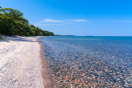 A serene beach with smooth pebbles and clear water under a bright blue sky, bordered by lush green trees.
