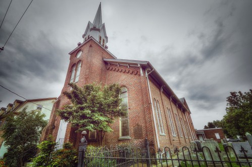 A brick church with a tall steeple, surrounded by trees and a wrought iron fence, under a cloudy sky.