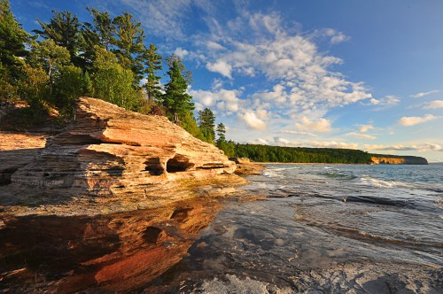 Rocky shoreline with reflections in water, surrounded by trees and a blue sky with scattered clouds.
