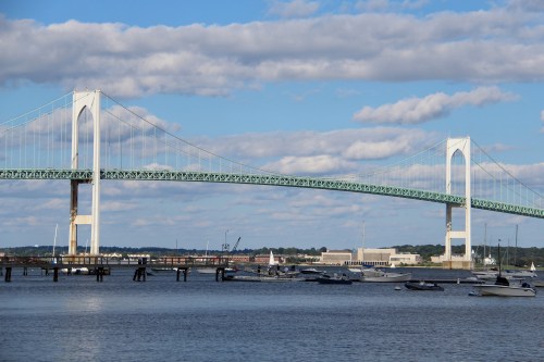 A suspension bridge spans across a calm waterway, with boats moored nearby and clouds in a blue sky.