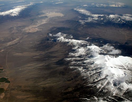 Aerial view of snow-capped mountains with valleys and plains below, showcasing a rugged, natural landscape.