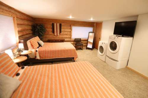 Cozy bedroom with two beds, a TV, and a laundry area, featuring wooden walls and warm orange bedding.