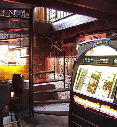 A vintage jukebox stands near a spiral staircase in a dimly lit bar with exposed brick walls.