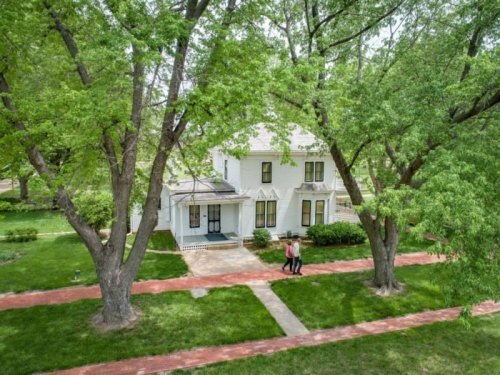 Aerial view of a white house surrounded by green trees, with two people walking on a brick path in the foreground.