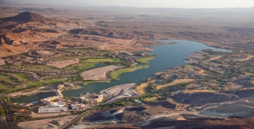 Aerial view of a desert landscape with a lake, golf courses, and residential areas surrounded by mountains.