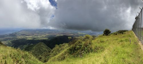 Panoramic view of lush green mountains and valleys under a cloudy sky, with a coastline visible in the distance.