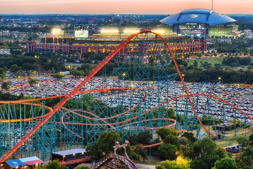 A vibrant amusement park with a roller coaster in the foreground and a stadium in the background at sunset.