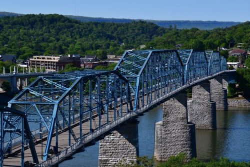 A blue steel bridge spans a river, surrounded by green hills and buildings under a clear blue sky.