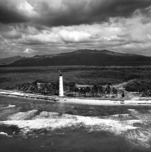 A black and white aerial view of a lighthouse on a coastal landscape with waves and mountains in the background.