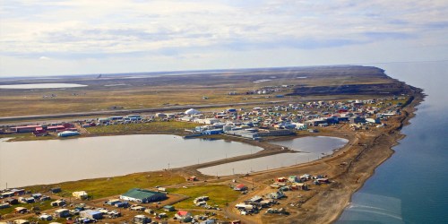 Aerial view of a small coastal town with colorful buildings, surrounded by water and open land under a cloudy sky.