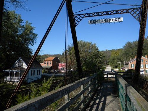 A wooden bridge leads into Bentonsport, featuring historic buildings and a clear blue sky.
