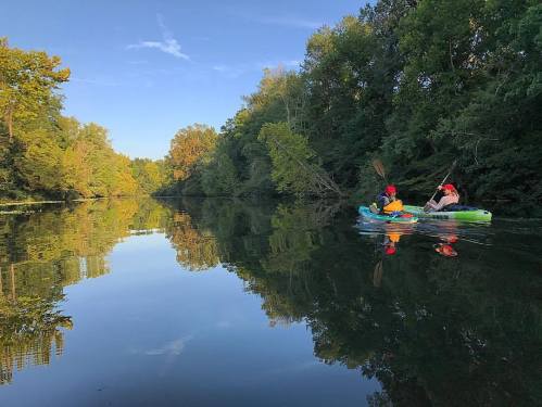 Two people kayak on a calm river surrounded by lush green trees, reflecting in the water under a clear blue sky.