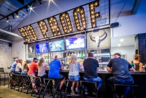 A lively bar scene with patrons seated at the counter, illuminated by a "PROOF" sign and multiple TVs in the background.