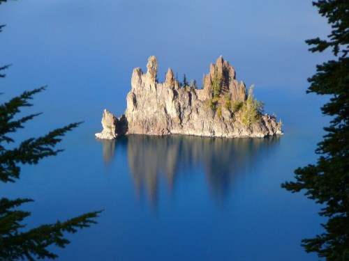 A rocky island with trees reflected in calm blue water, surrounded by a serene landscape.