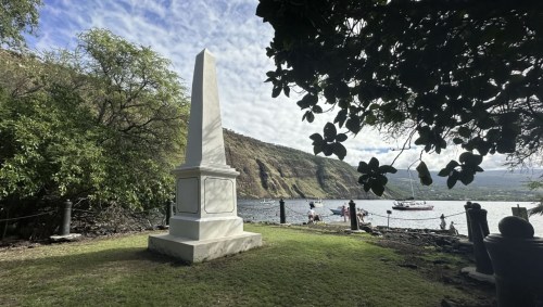 A white obelisk monument stands on green grass by a calm bay, with boats and hills in the background under a cloudy sky.