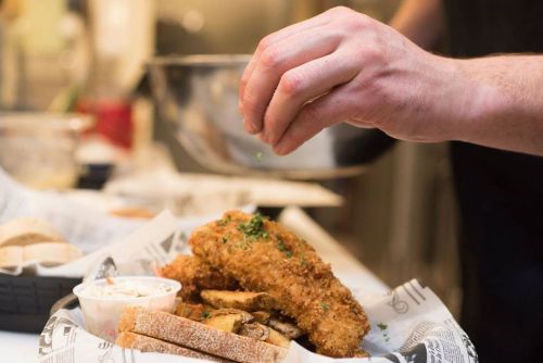 A hand sprinkles seasoning over a plate of fried food, including fish and fries, in a kitchen setting.